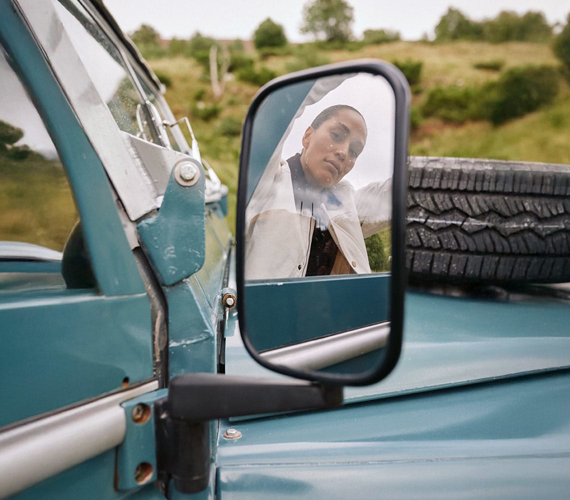 A woman is seen in the side mirror of a car, her reflection framed by the mirror's edge and a distant landscape.